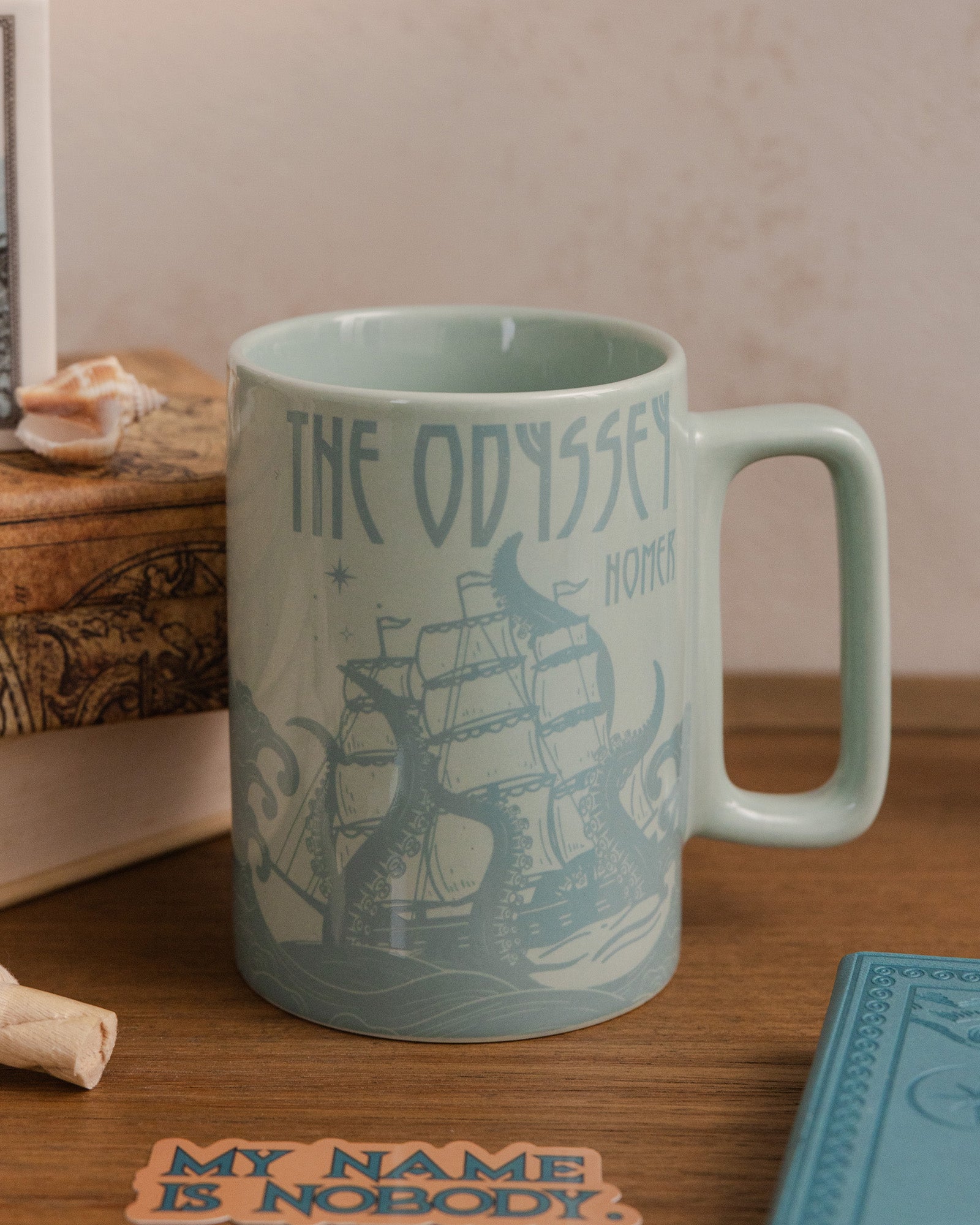 Mug with 'The Odyssey' design on a wooden surface with books and a shell.