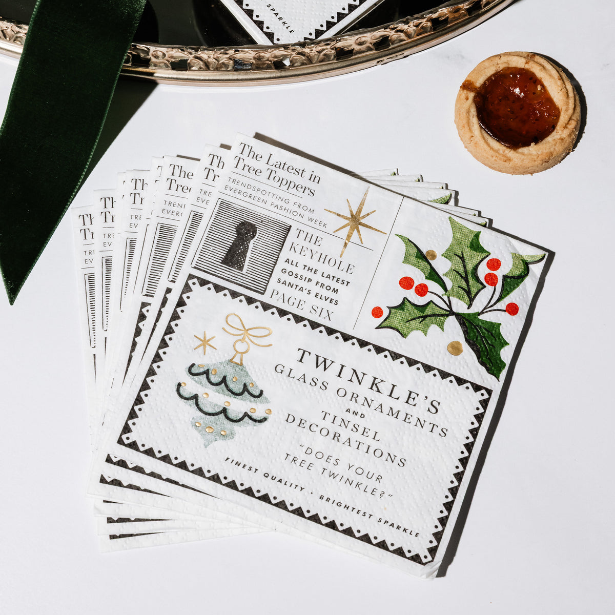 square paper napkins from The Christmas Chronicle, featuring vintage holiday illustrations, text snippets, and festive designs on a white table surrounded by holiday items