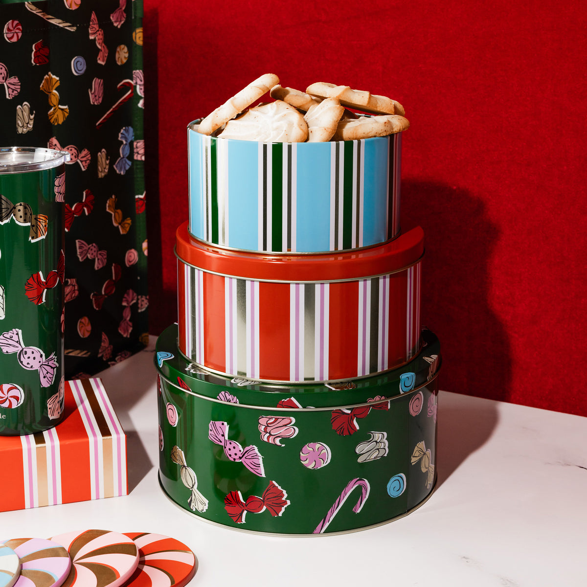 Three stacked round metal tins with festive holiday candy patterns and striped accents, with cookies visible in the top tin, set on a white surface against a red background.