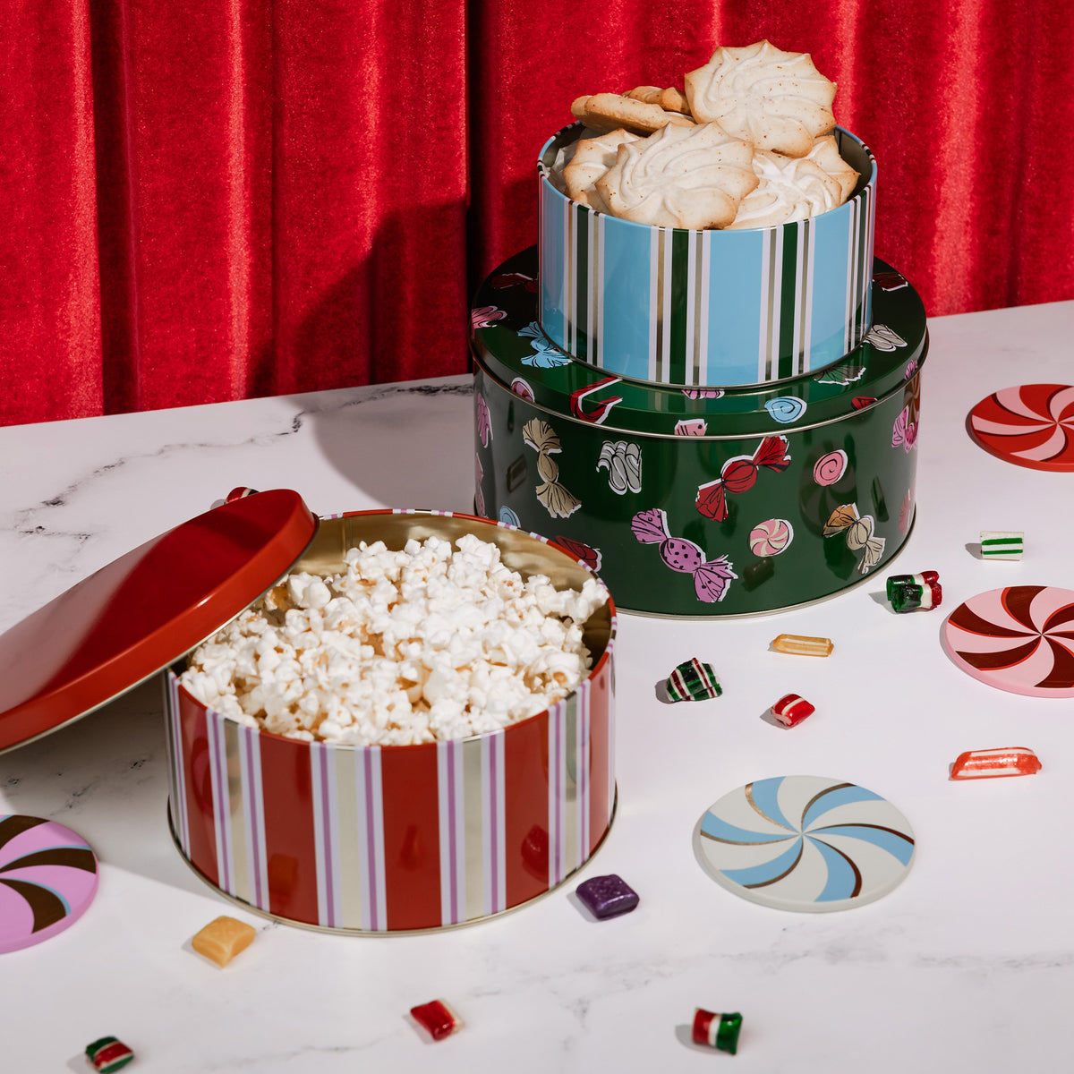 Three decorative holiday metal tins filled with snacks and cookies, arranged on a white marble surface with scattered candy pieces and a red curtain backdrop.
