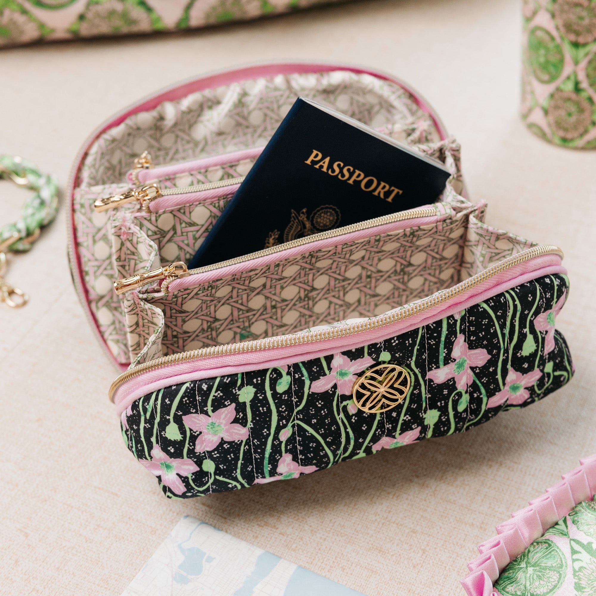 A patterned zippered pouch with pink trim and a gold logo emblem, shown open with a passport inside on a beige fabric surface.