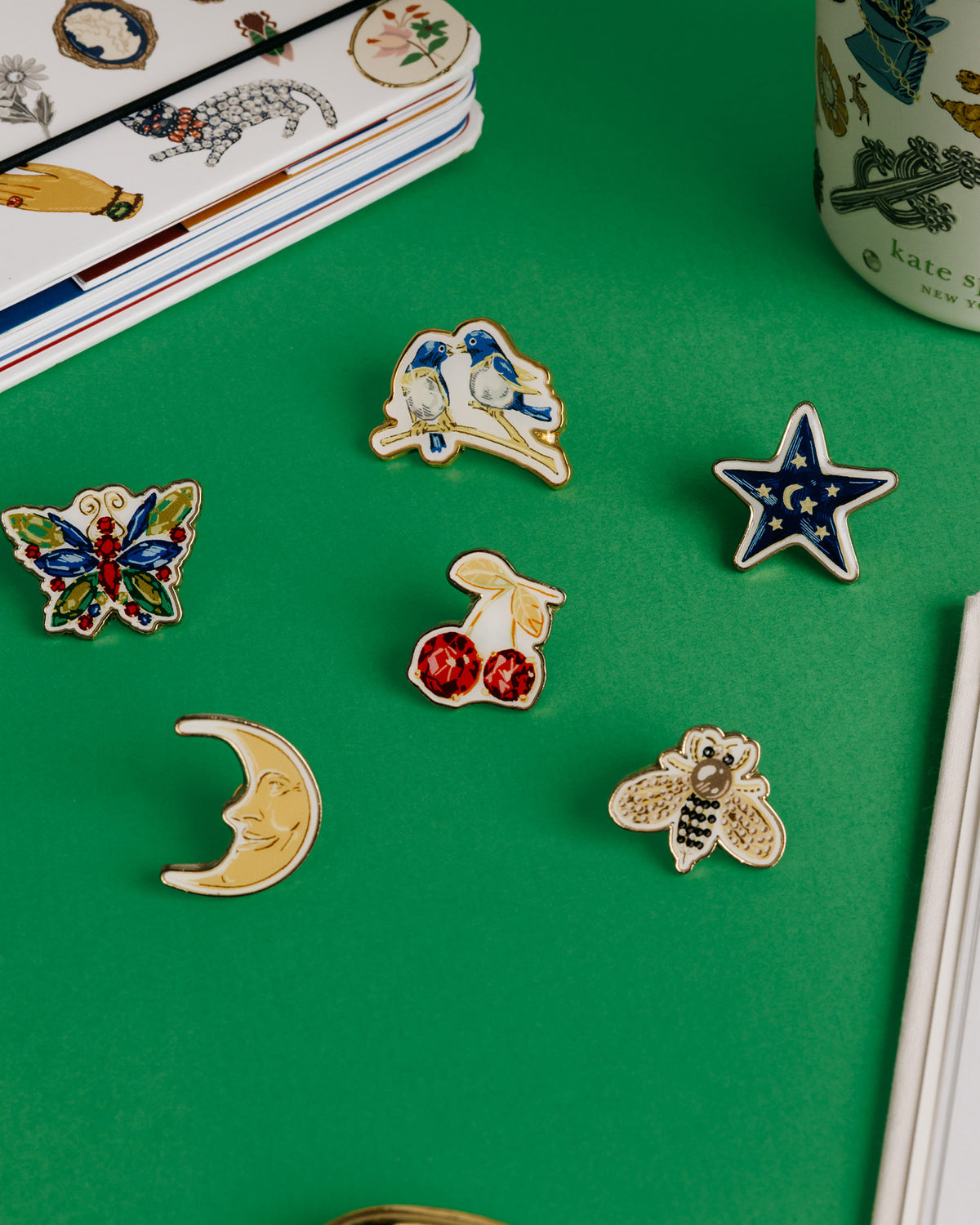 Kate Spade New York enamel pin set in a green presentation box, featuring six pins: cherries, a star, a bee, a crescent moon, a butterfly, and a bird on a green background