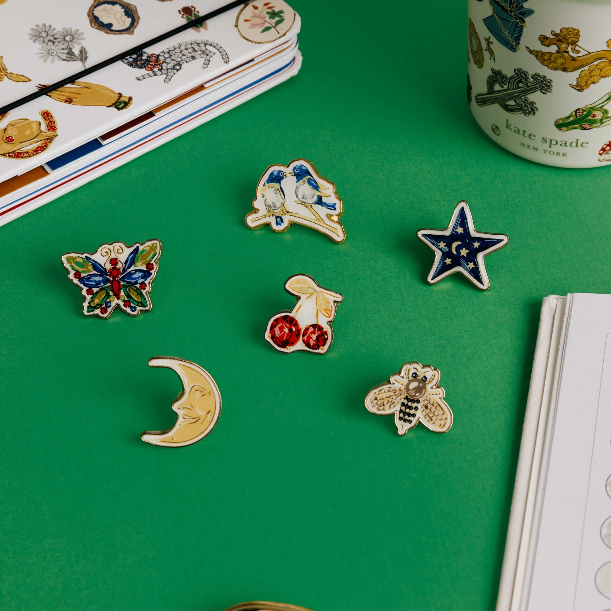 Kate Spade New York enamel pin set in a green presentation box, featuring six pins: cherries, a star, a bee, a crescent moon, a butterfly, and a bird on a green background