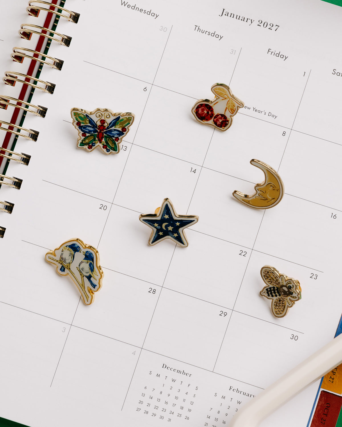 Kate Spade New York enamel pin set in a green presentation box, featuring six pins: cherries, a star, a bee, a crescent moon, a butterfly, and a bird on a planner