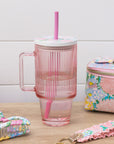 Pink striped mug with straw, floral pouch, and hair accessories on a wooden surface.