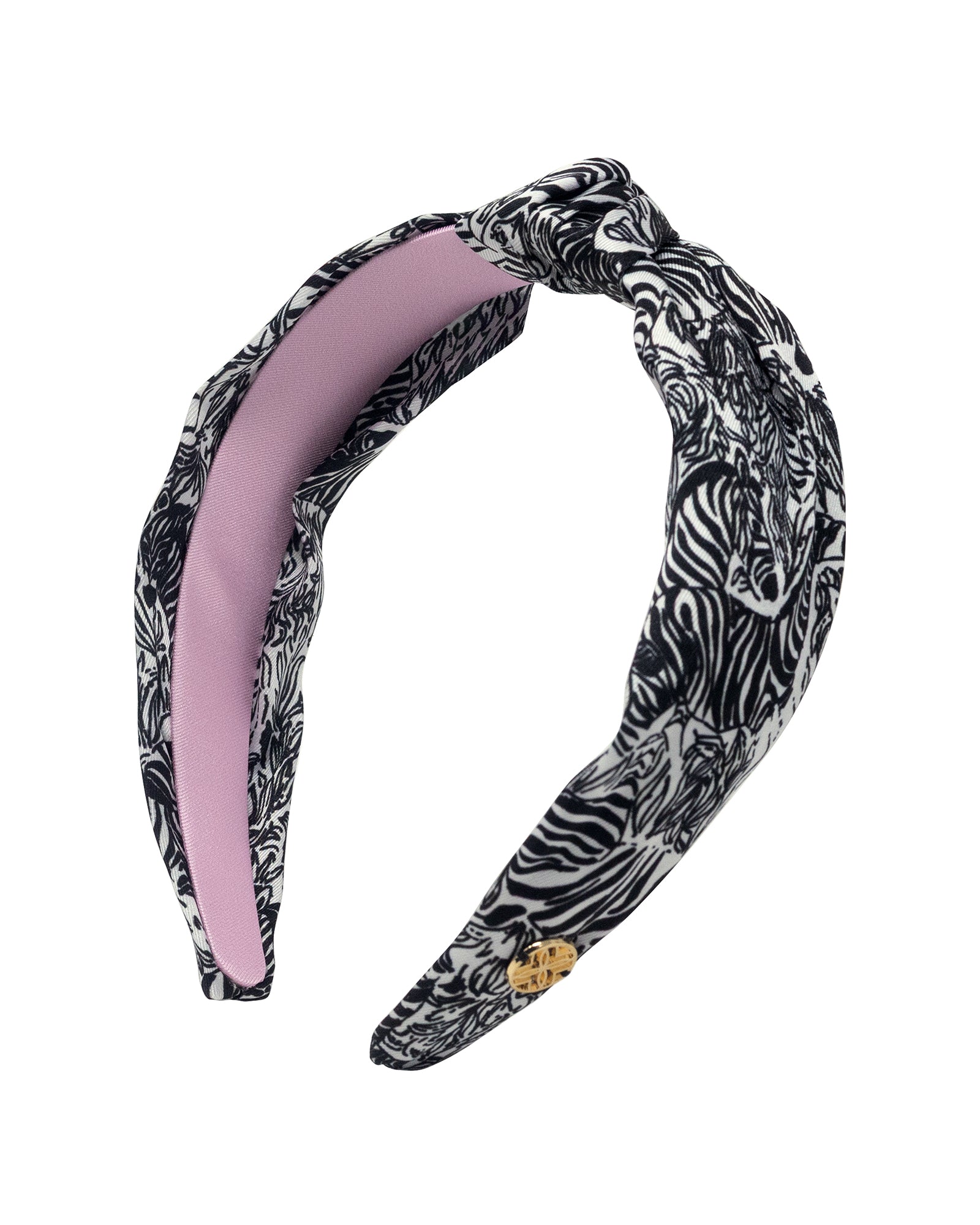 A twisted fabric headband featuring a black and white zebra print on the exterior and a solid light pink interior, with a small gold logo charm.