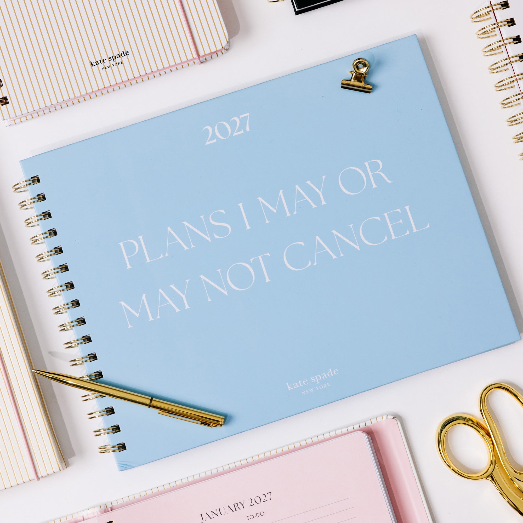 A light blue spiral-bound 2027 planner titled 'PLANS I MAY OR MAY NOT CANCEL' in white lettering, styled on a white desk with gold scissors, a gold pen, and other notebooks.