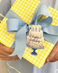 Gift wrapped in yellow checkered paper with a blue ribbon and 'Happy Birthday' cake tag.