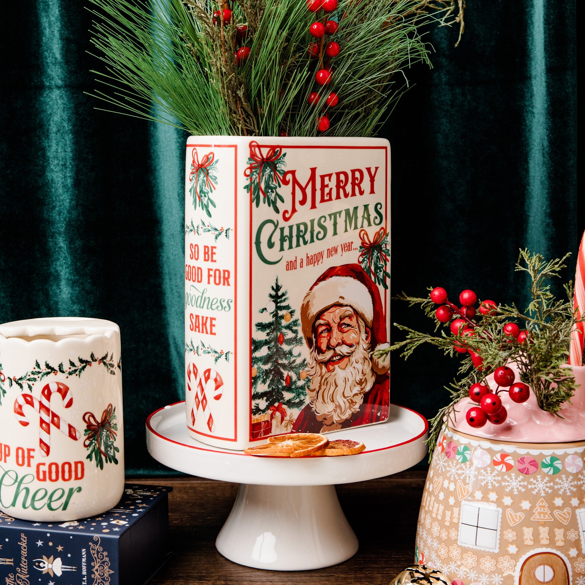 A vintage-style ceramic Christmas canister with a Santa Claus illustration and 'Merry Christmas' text, displayed on a white pedestal stand. The canister is decorated with red berries, pine branches, and holiday greenery, set against a dark green velvet curtain background.