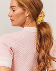 Woman with long hair tied in a ponytail with a yellow scrunchie, wearing a pink cable knit sweater.
