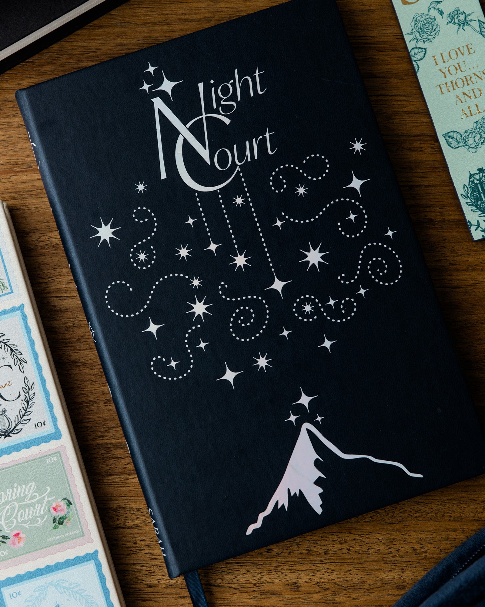 Black notebook with white star design and 'Night Court' text on a wooden surface