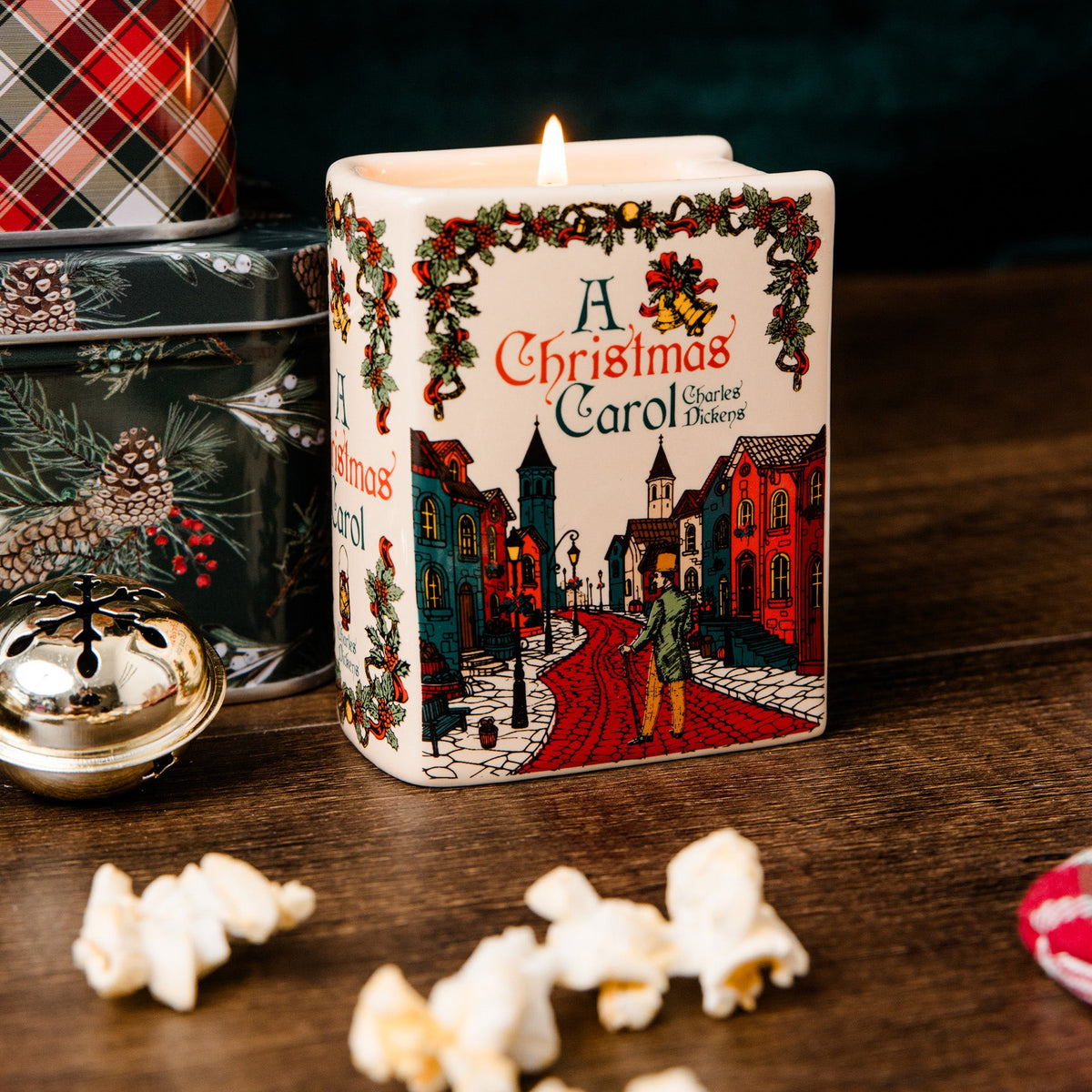 A square white candle with a lit flame, printed with a vintage-style 'A Christmas Carol' street scene and holiday garland artwork, displayed on a wooden surface with popcorn and holiday-themed containers nearby.
