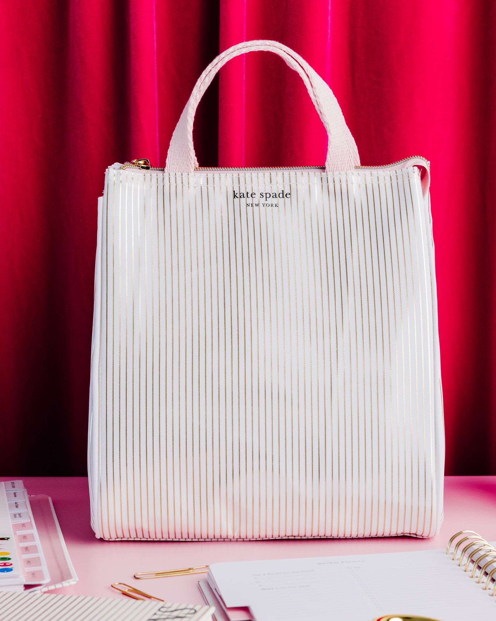 A Kate Spade New York lunch bag in a textured, vertically pleated off-white fabric, set against a vibrant pink and red curtain backdrop with stationery props on a pink table.
