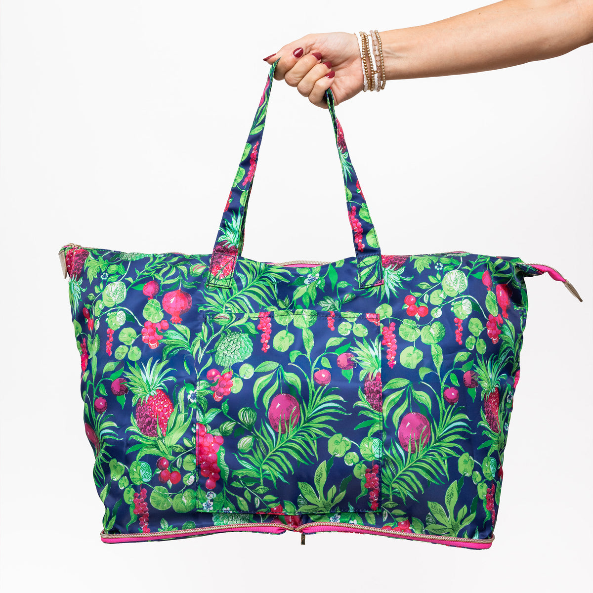 model holding a large tote bag with a vibrant tropical fruit and leaf print on a navy blue background, featuring bright pink accents along the zipper and handle edges.