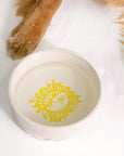 White ceramic bowl with yellow sun design on a white background, with a dog's paw partially visible.
