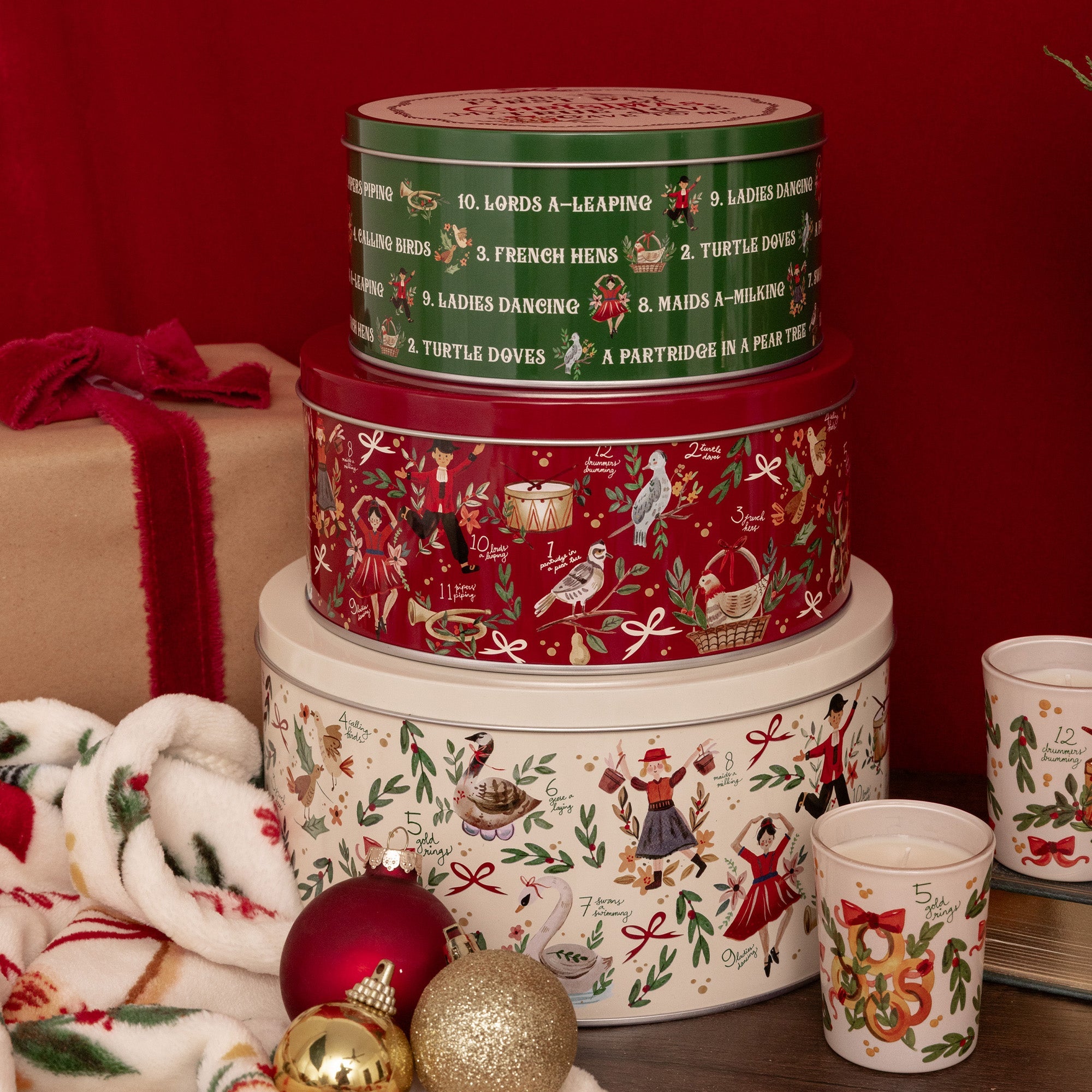 Three stacked round Christmas-themed metal tins with matching holiday cups, alongside a wrapped gift box and ornaments, set against a dark red background.