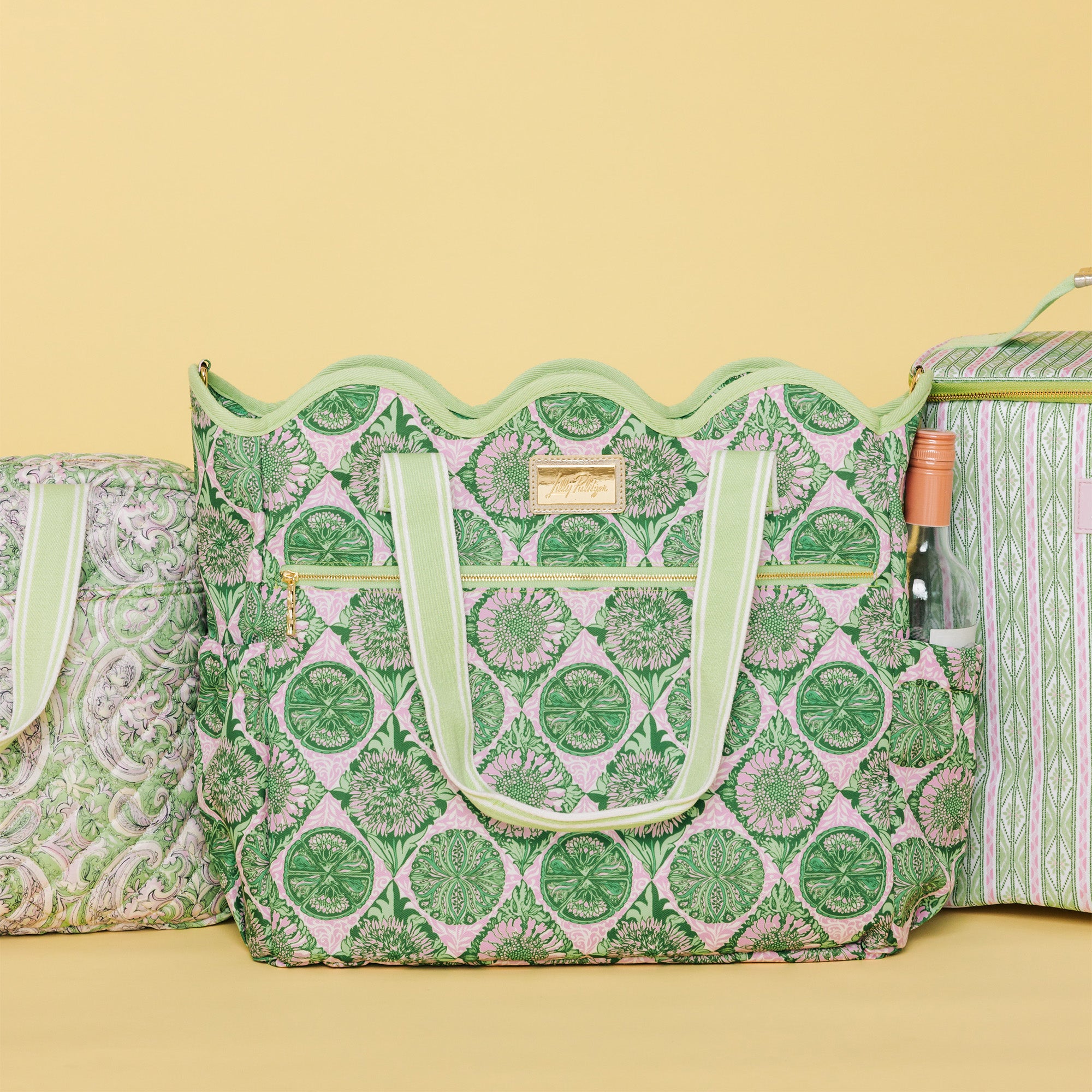 A green and pink patterned tote bag with scalloped edges and light green handles, shown alongside matching patterned bags on a pale yellow background.