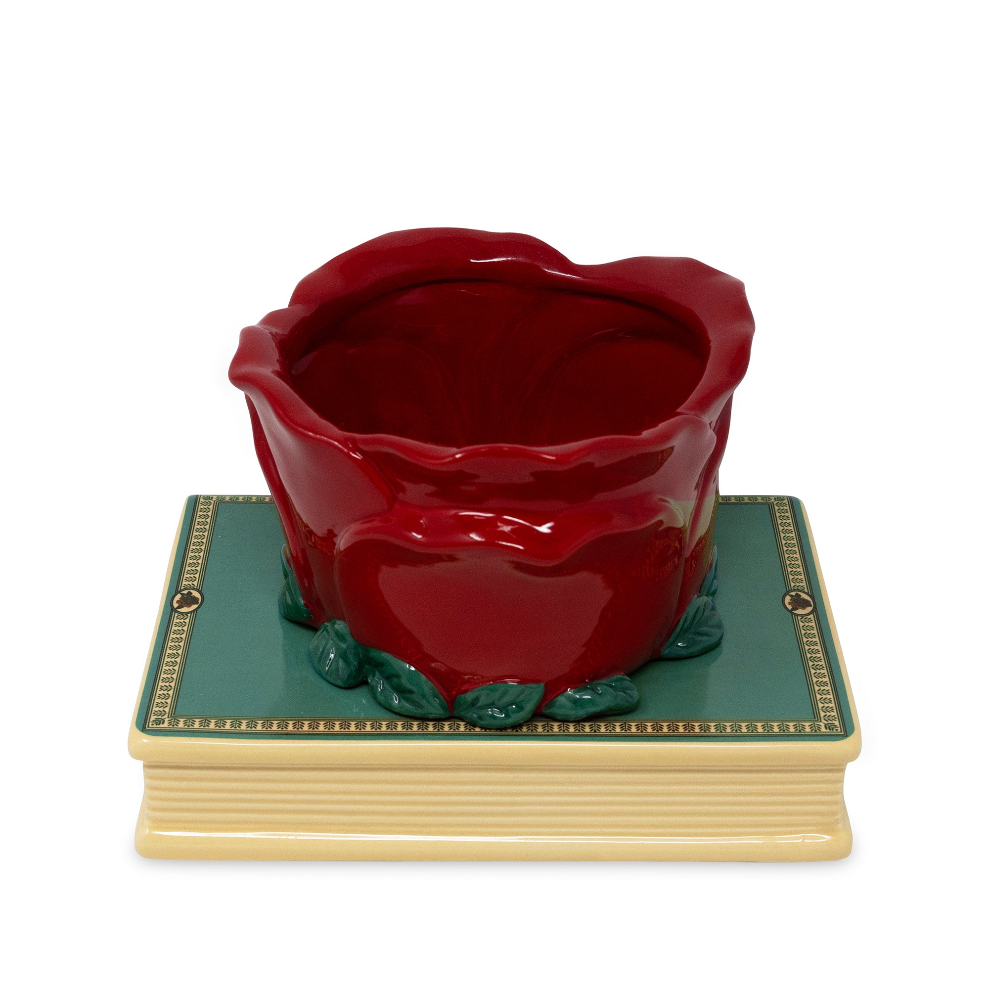 A glossy red ceramic rose-shaped container or planter displayed on top of a decorative book-shaped base with a teal cover and cream pages, set against a plain white background.