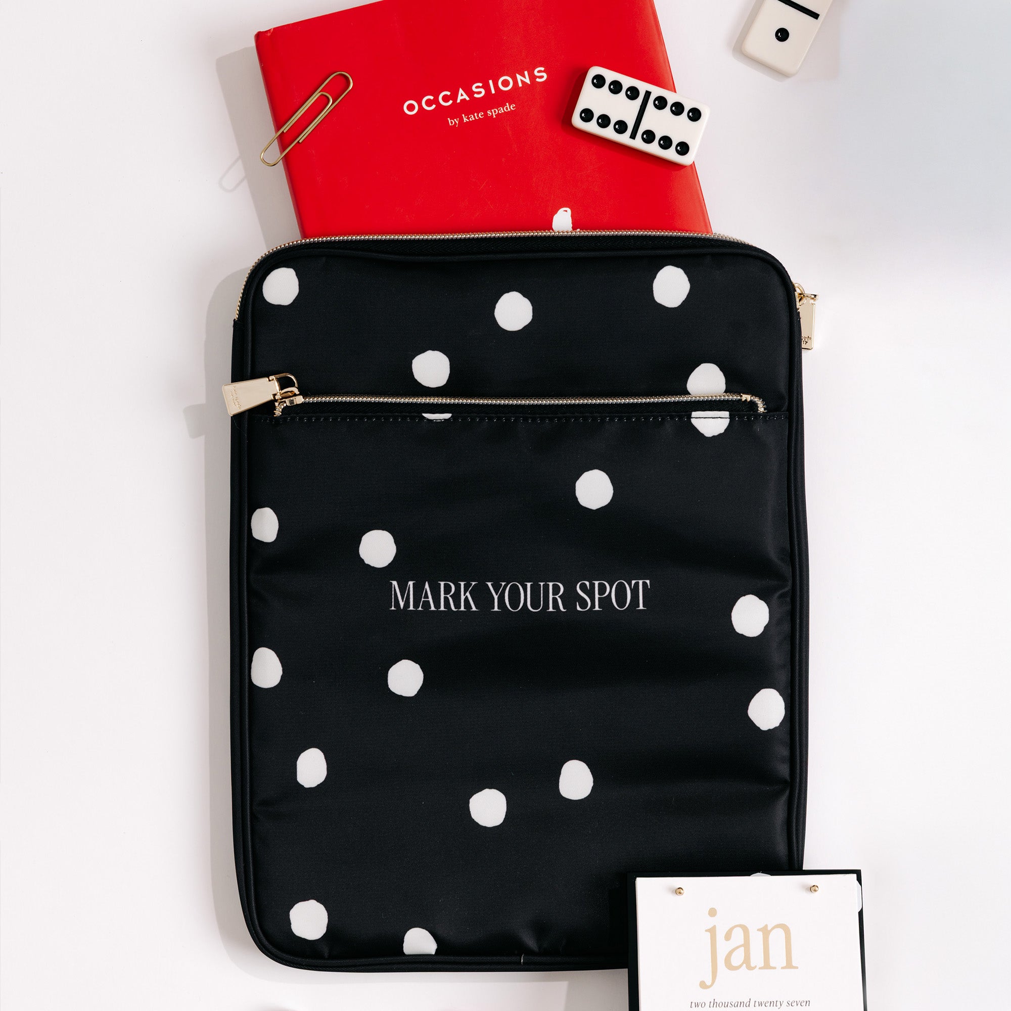 A black nylon tablet sleeve with white polka dots and the words 'MARK YOUR SPOT' printed in white, styled on a white background with a red notebook, paperclip, dice, and a hangtag.