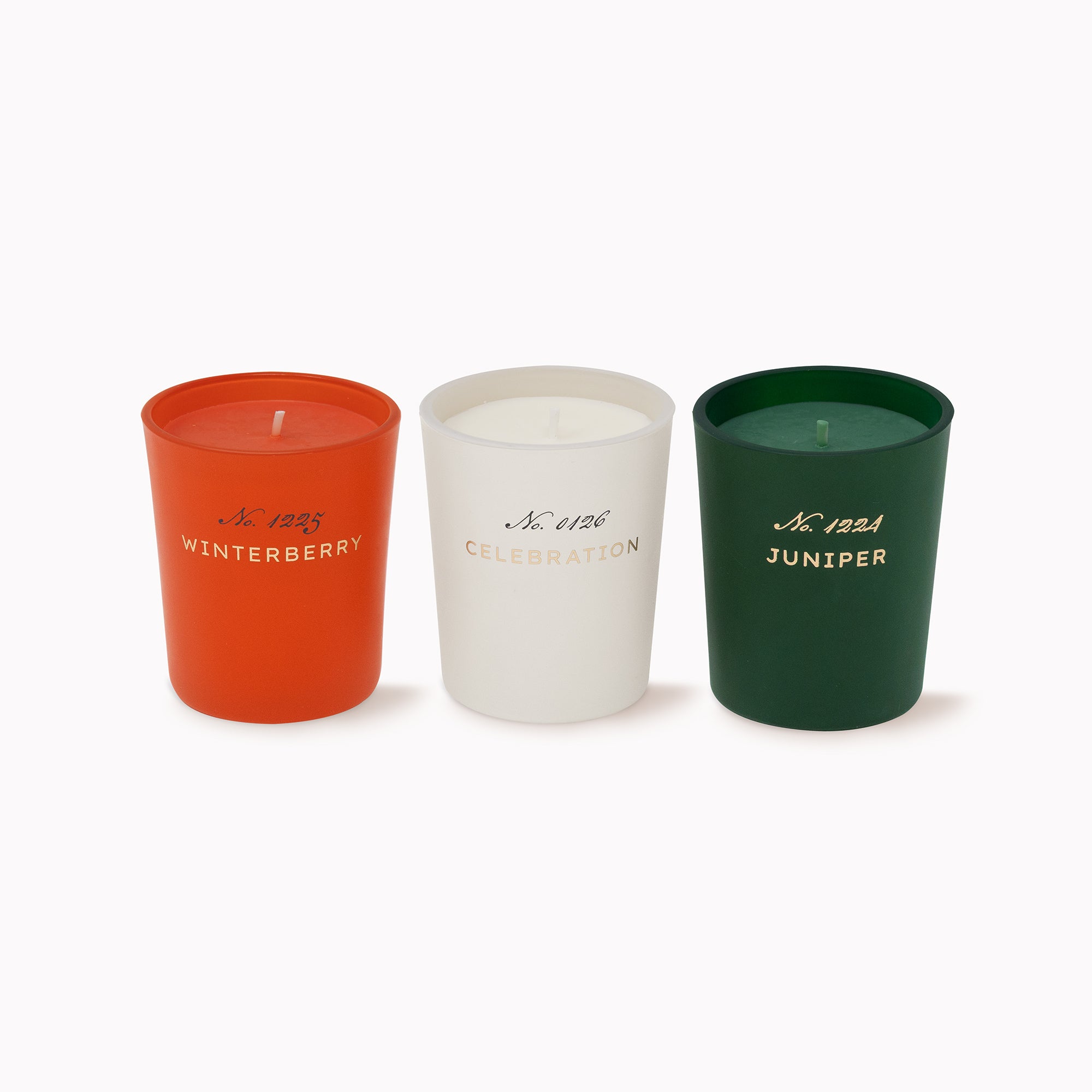 Three scented candles in matte-colored glass candles on a white background: a burnt orange candle labeled Winterberry, a white candle labeled Celebration, and a dark green candle labeled Juniper.