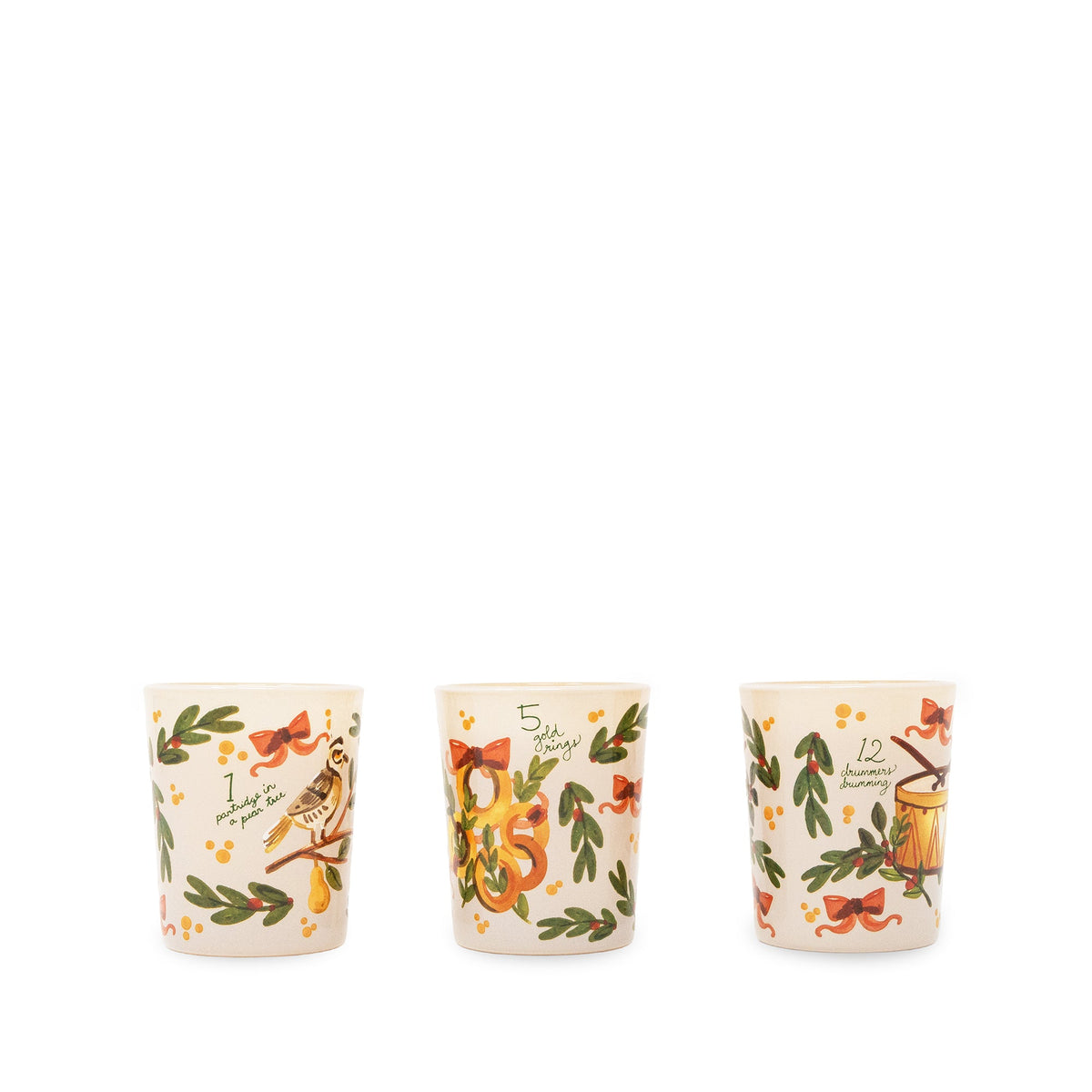 Three small ceramic candles with a warm cream background, decorated with green leaves, red bows, small yellow flowers, and illustrated holiday motifs including a bird, a nut, and a mug, shown on a plain white background.