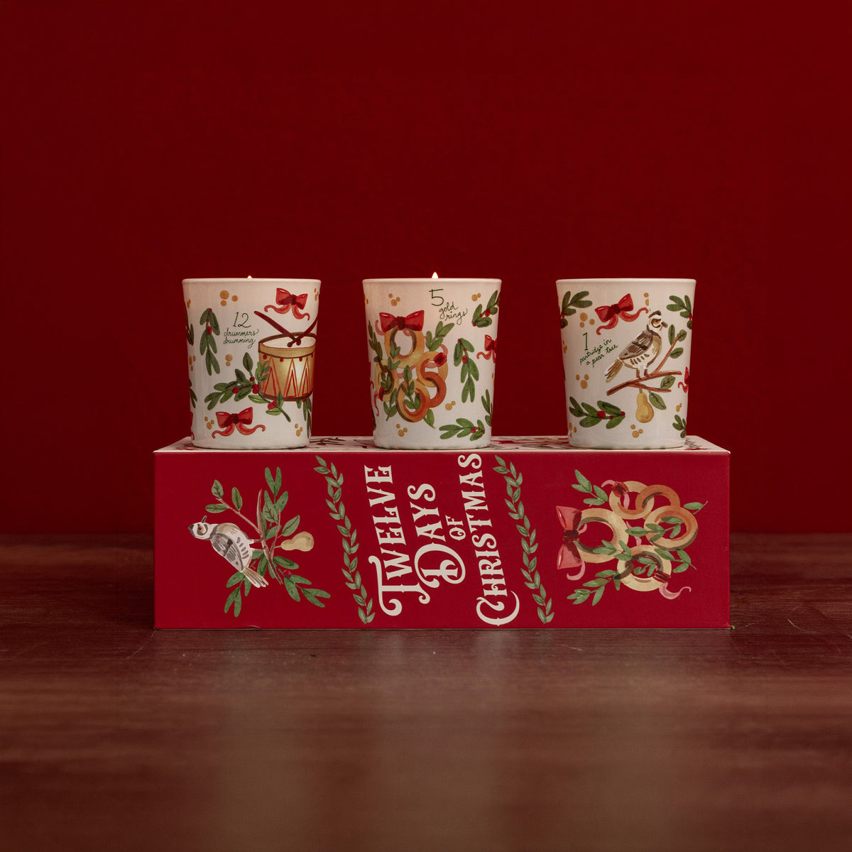 Three white Christmas-themed candles with festive illustrations, displayed on top of a red gift box printed with 'Twelve Days of Christmas' against a dark red background.