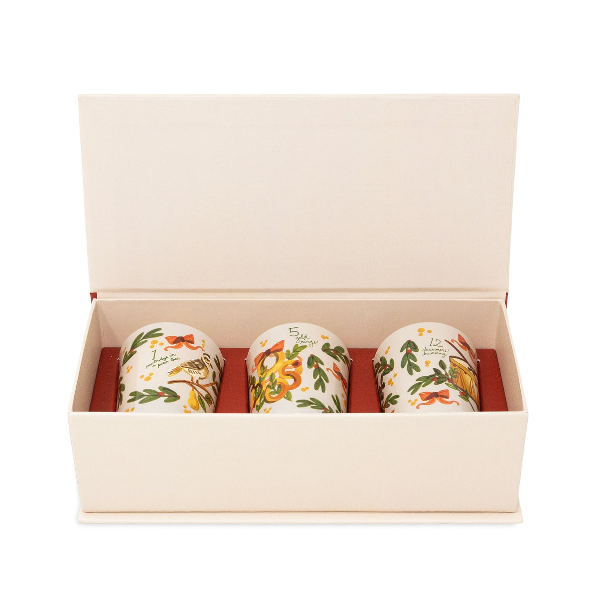 Three ceramic mugs with holiday illustrations displayed inside an open cream-colored gift box with a red interior.