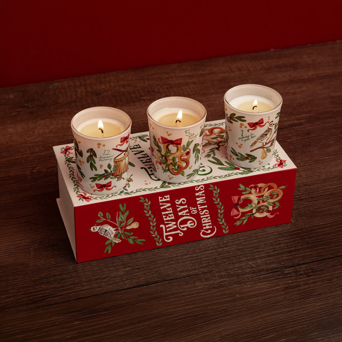 Three lit holiday candles in white glass holders with red and green Christmas illustrations, displayed on top of a red gift box printed with 'Three Days of Christmas' on a dark wooden surface.