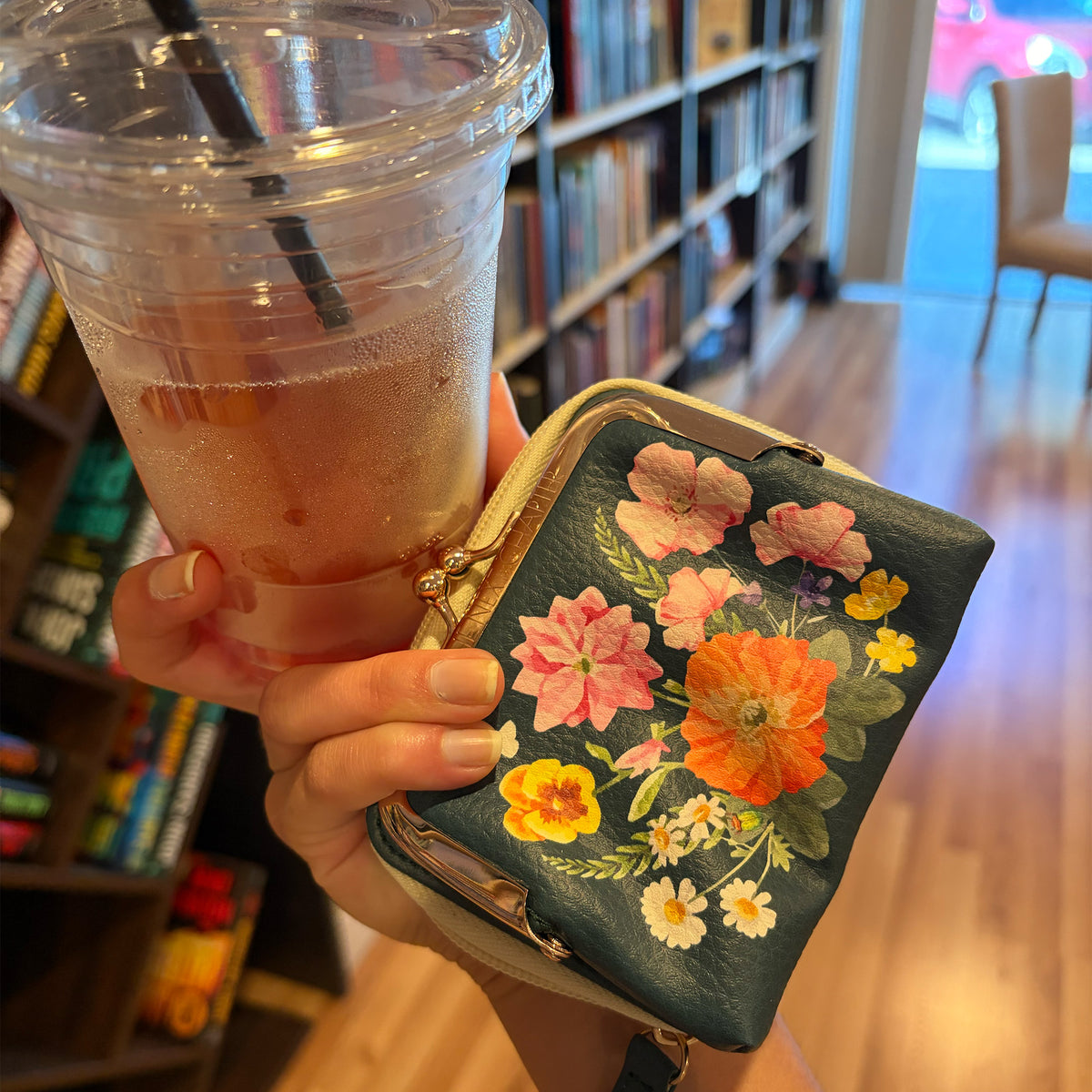 A hand holds a small floral coin purse next to a plastic iced coffee cup in a bookstore or library setting with bookshelves in the background.