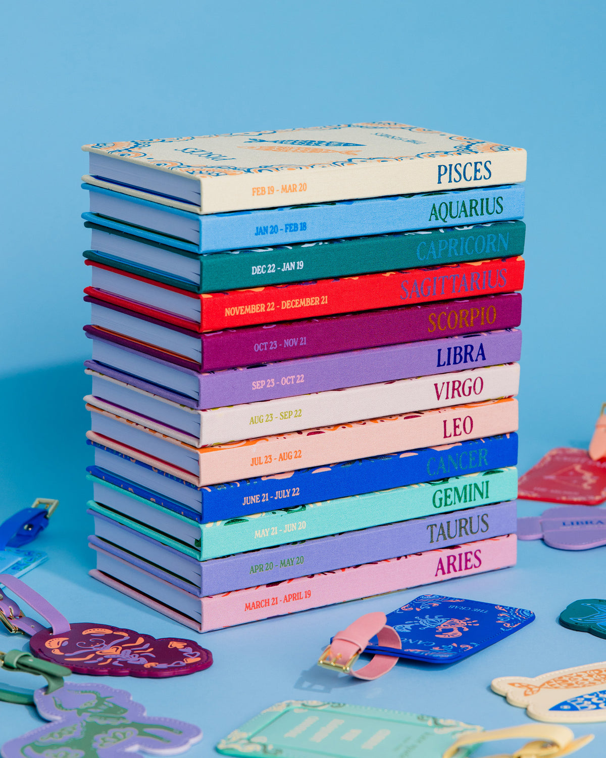 A stack of colorful zodiac-themed notebooks or journals with pastel covers and printed zodiac names, arranged on a light blue surface with matching zodiac-shaped accessories scattered around.