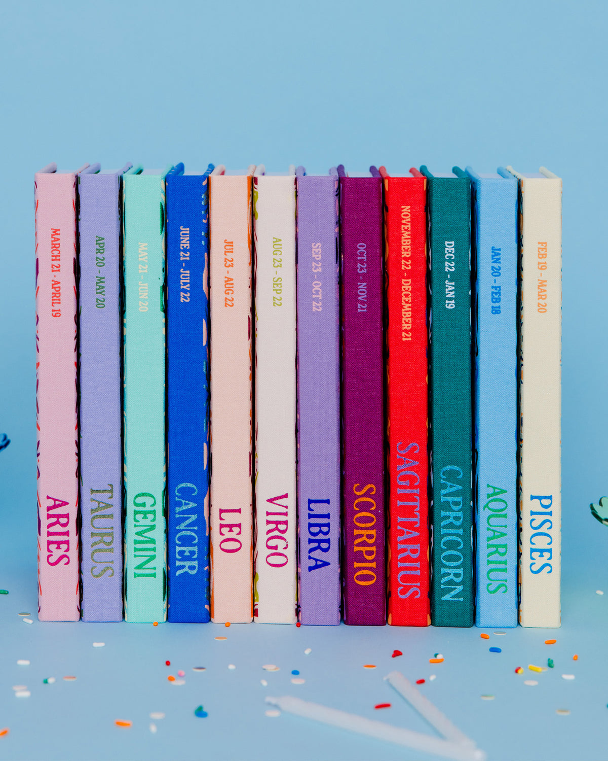 A set of twelve hardcover books arranged in a row, each with a different colored spine representing a zodiac sign, set against a light blue background with decorative cupcakes on the sides.