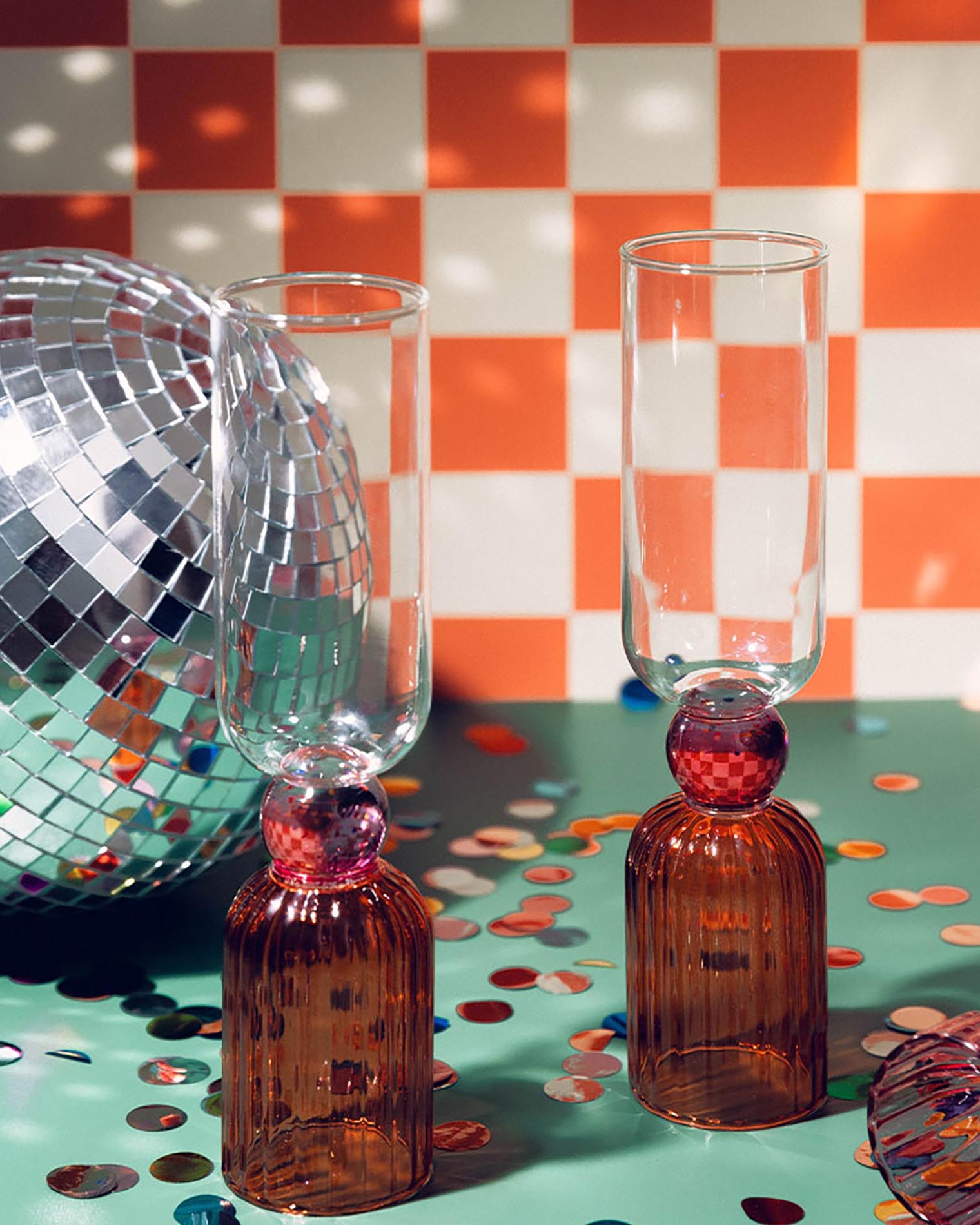 Two amber glass bottles with red stoppers and clear glasses on a checkered tablecloth with disco ball and confetti.