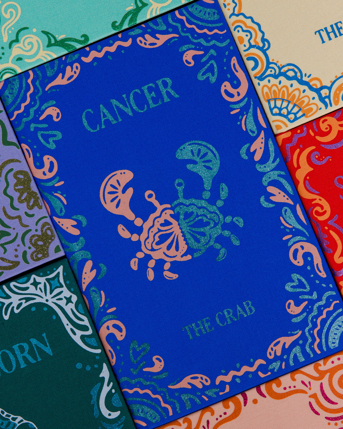 A set of colorful zodiac-themed cards or booklets arranged in a flat lay. The central card is blue with a teal crab illustration and the word 'CANCER'.