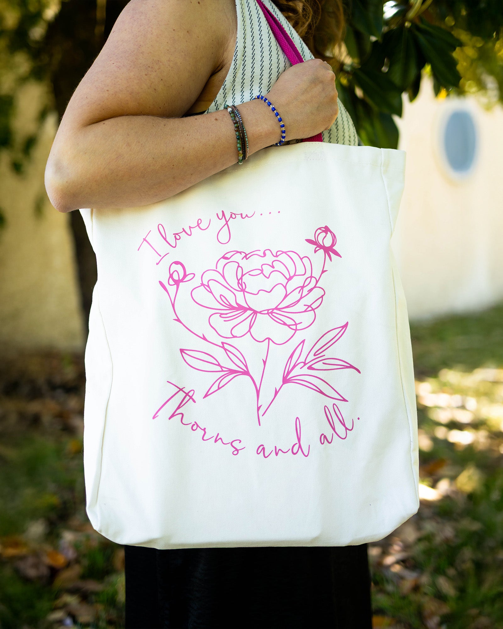 model carrying white tote with pink strap and lettering that says 'I love you...thorns and all' with a sketched floral print