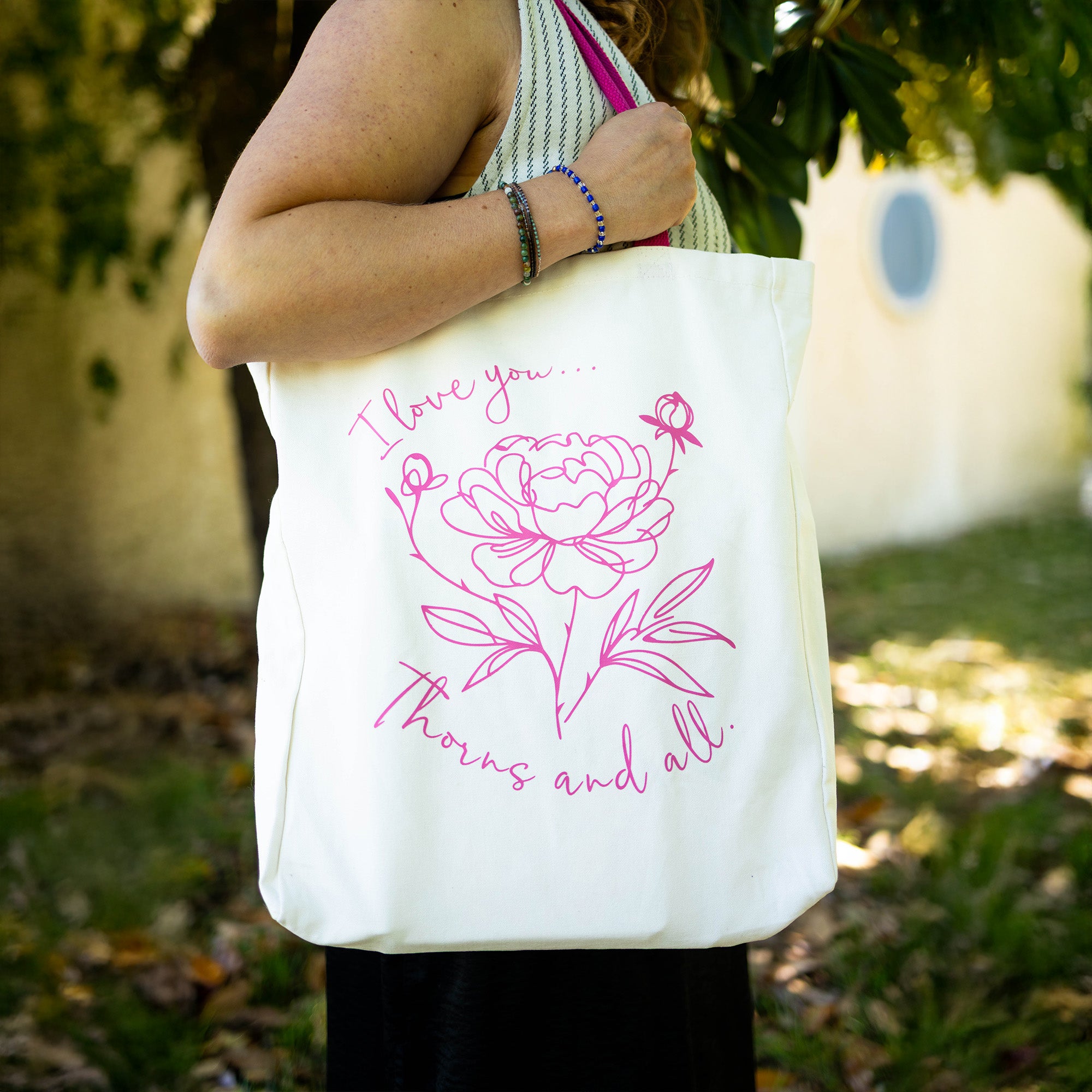 model carrying white tote with pink strap and lettering that says 'I love you...thorns and all' with a sketched floral print