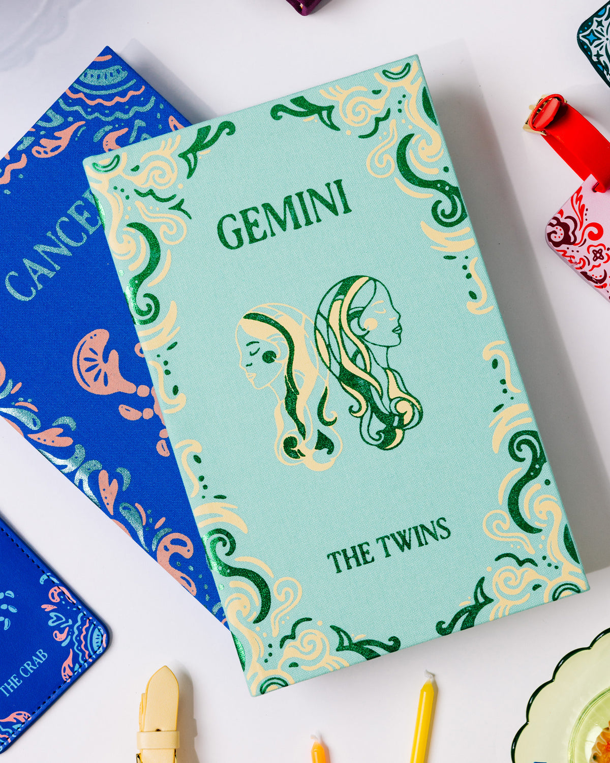A mint green Gemini zodiac-themed notebook or card case with green and cream swirl patterns and twin illustrations, styled on a white surface with other colorful astrology-themed accessories.