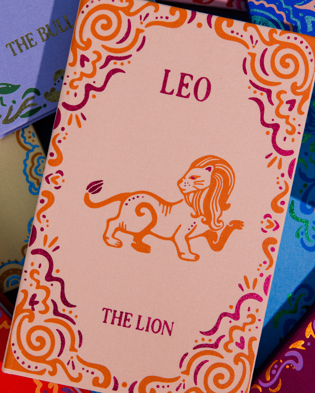 A close-up of a pink zodiac card titled 'LEO THE LION' with an orange lion illustration and decorative swirl borders, surrounded by other colorful patterned cards.