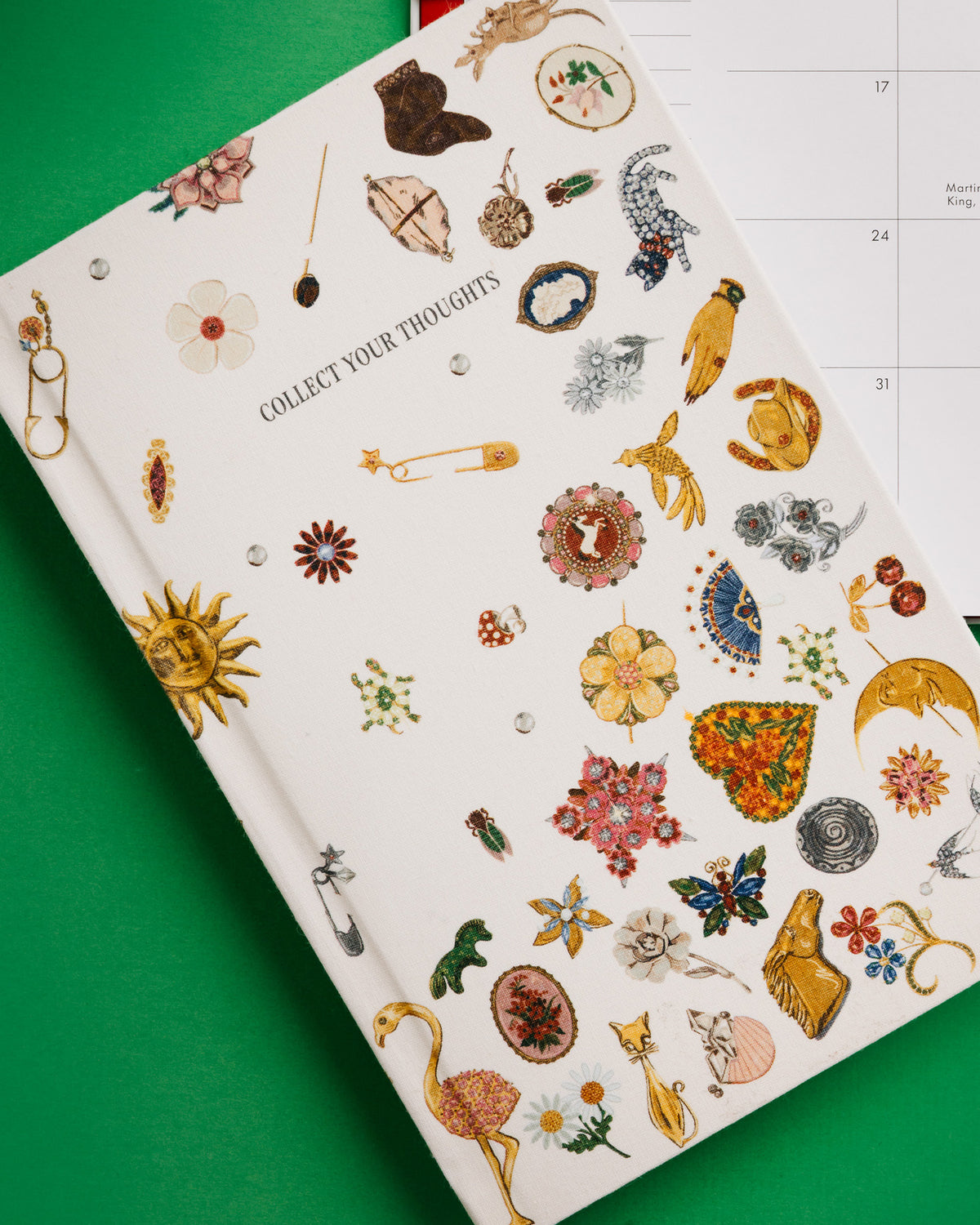 A cream-colored hardcover journal titled 'COLLECT YOUR THOUGHTS' decorated with various colorful vintage-style illustrations of jewelry, flowers, and symbols, shown on a bright green background with a monthly calendar partially visible behind it.