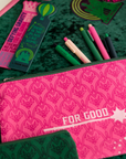 Pink pencil case with 'FOR GOOD' text on a green textured surface with stationery items.