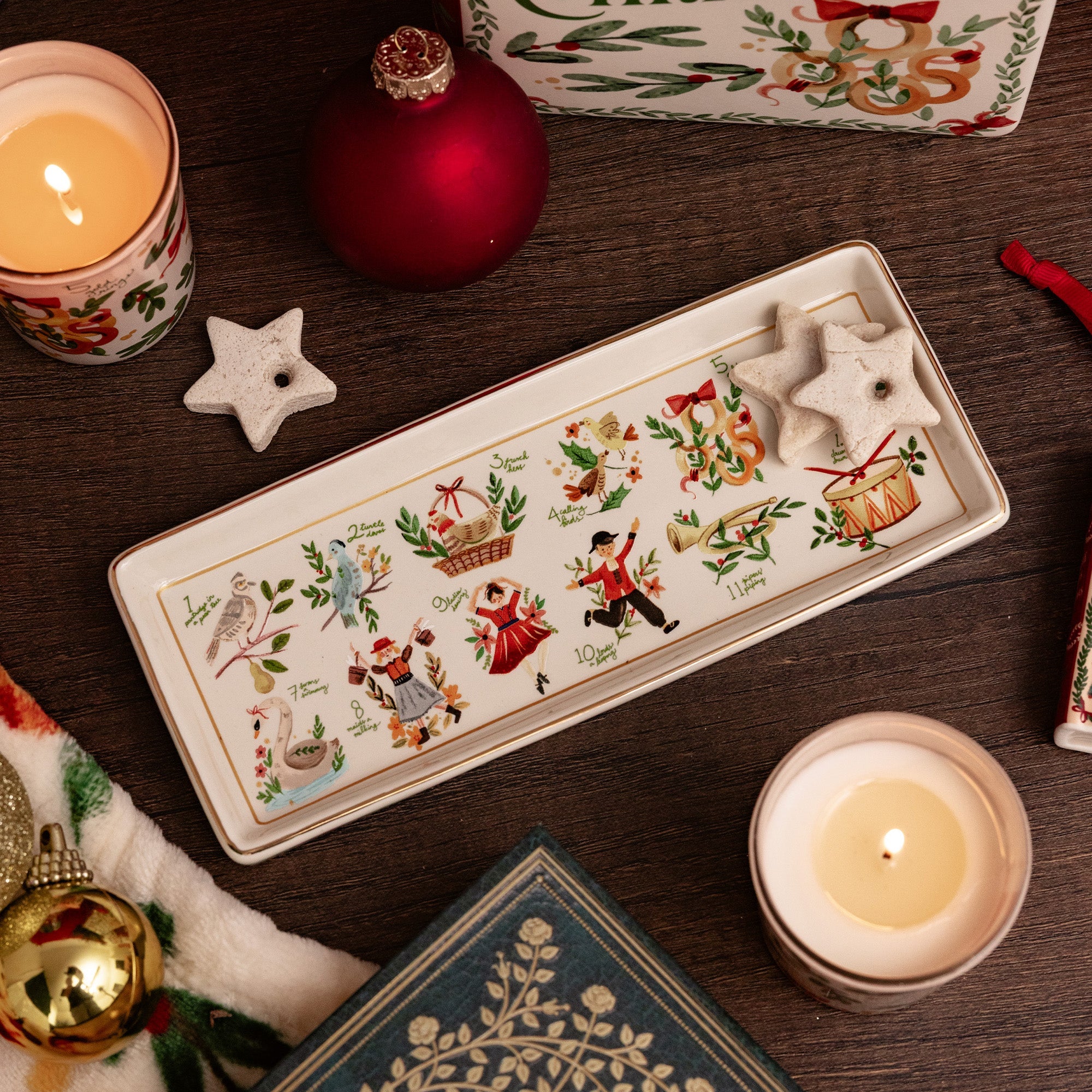 A rectangular ceramic Christmas tray with a cream background and colorful illustrated holiday motifs, styled on a dark wooden surface with candles, ornaments, a star candle holder, and a book.