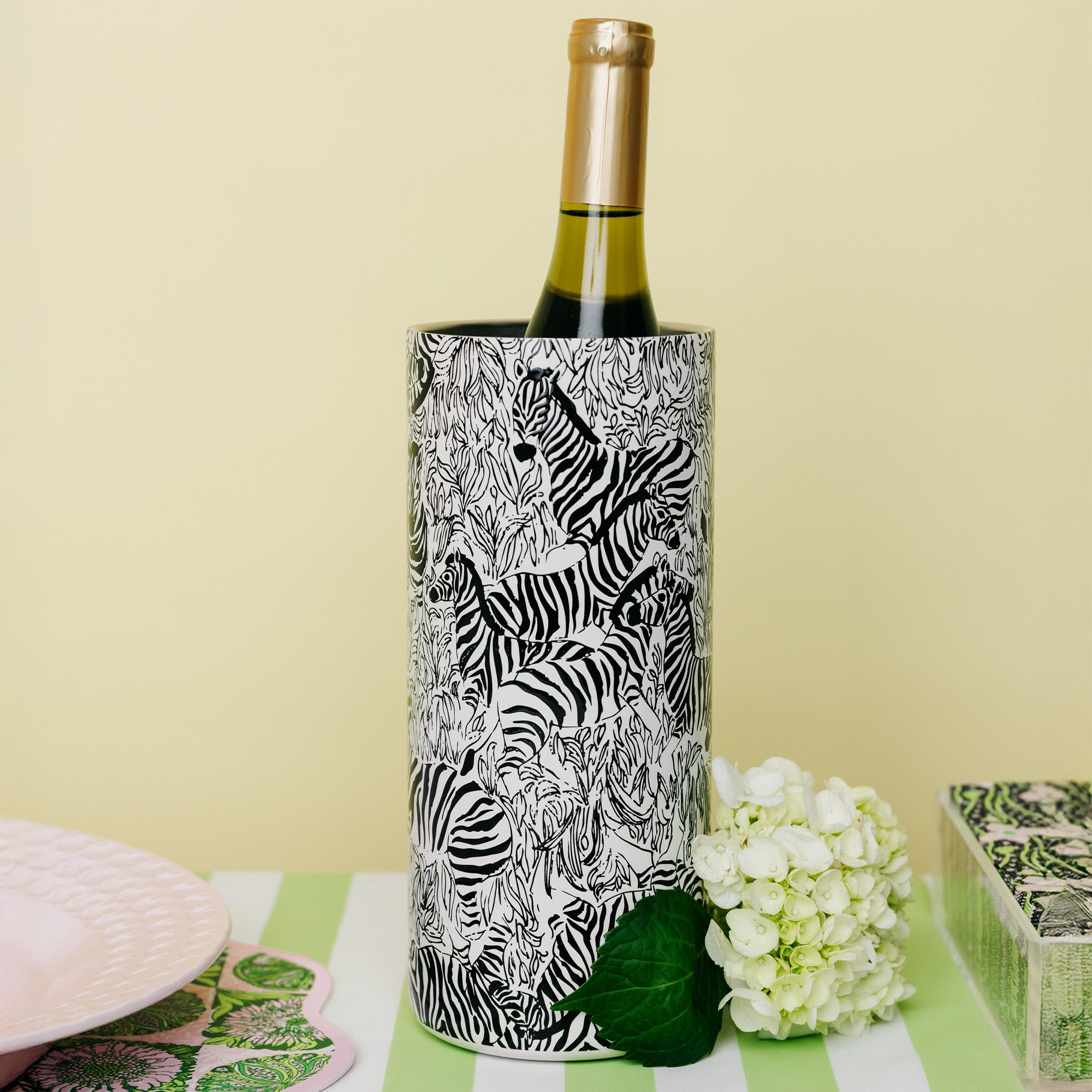 A wine bottle with a gold foil top stands inside a cylindrical holder covered in a black-and-white zebra print. The holder sits on a green-and-white striped surface with a pale yellow background, white flowers, and partial decorative objects at the edges.