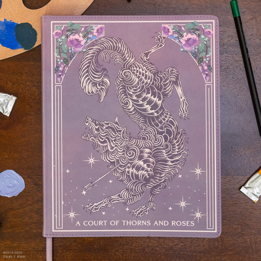 A purple hardcover journal titled 'A COURT OF THORNS AND ROSES' featuring a detailed line-art illustration of a goat-like creature, laid flat on a wooden desk with paint supplies nearby.