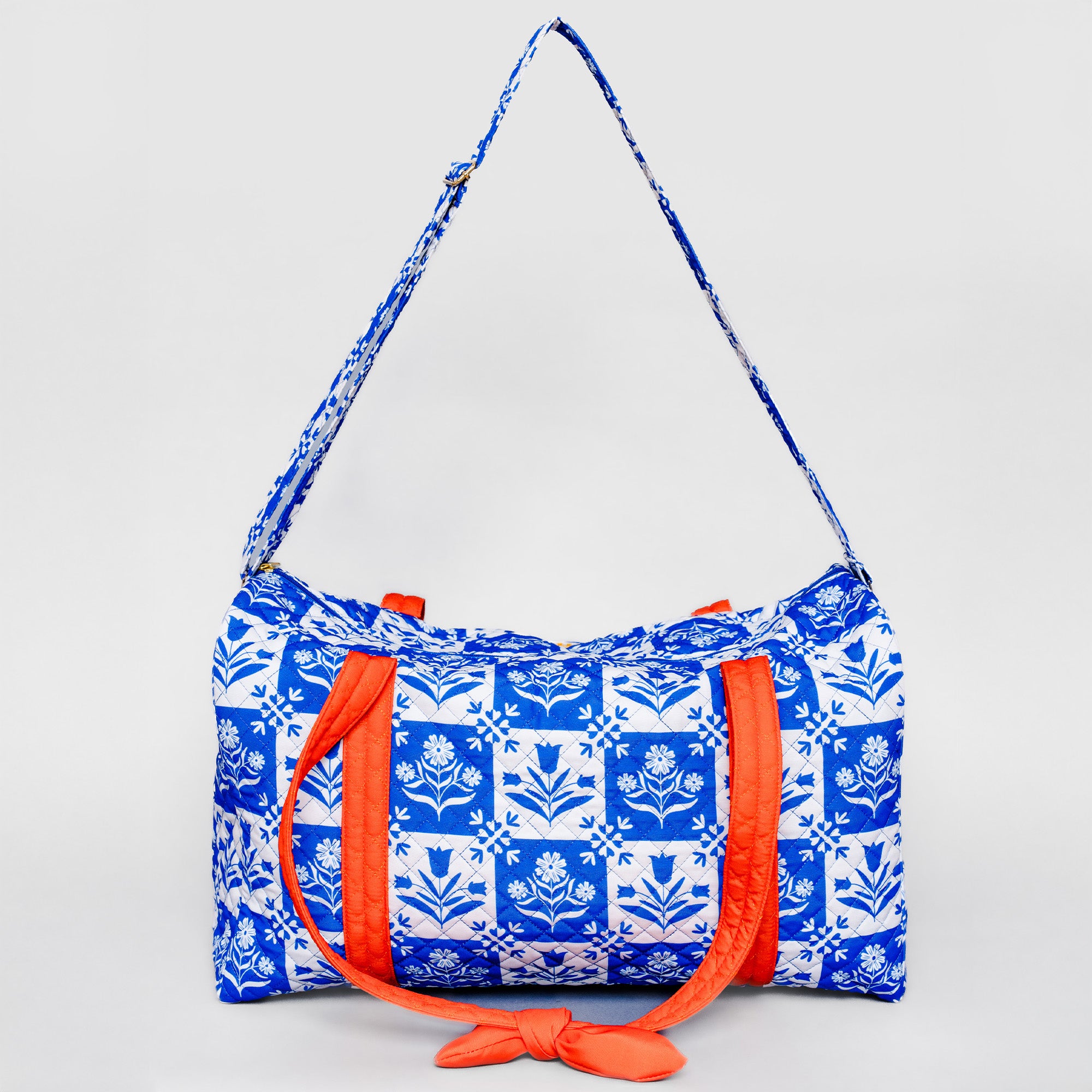 A blue and white patterned duffel bag with bright orange straps and a matching patterned shoulder strap, photographed against a light gray background.
