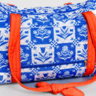 A quilted duffel bag featuring a blue and white floral block print pattern with bright orange straps and trim, shown against a light gray background.