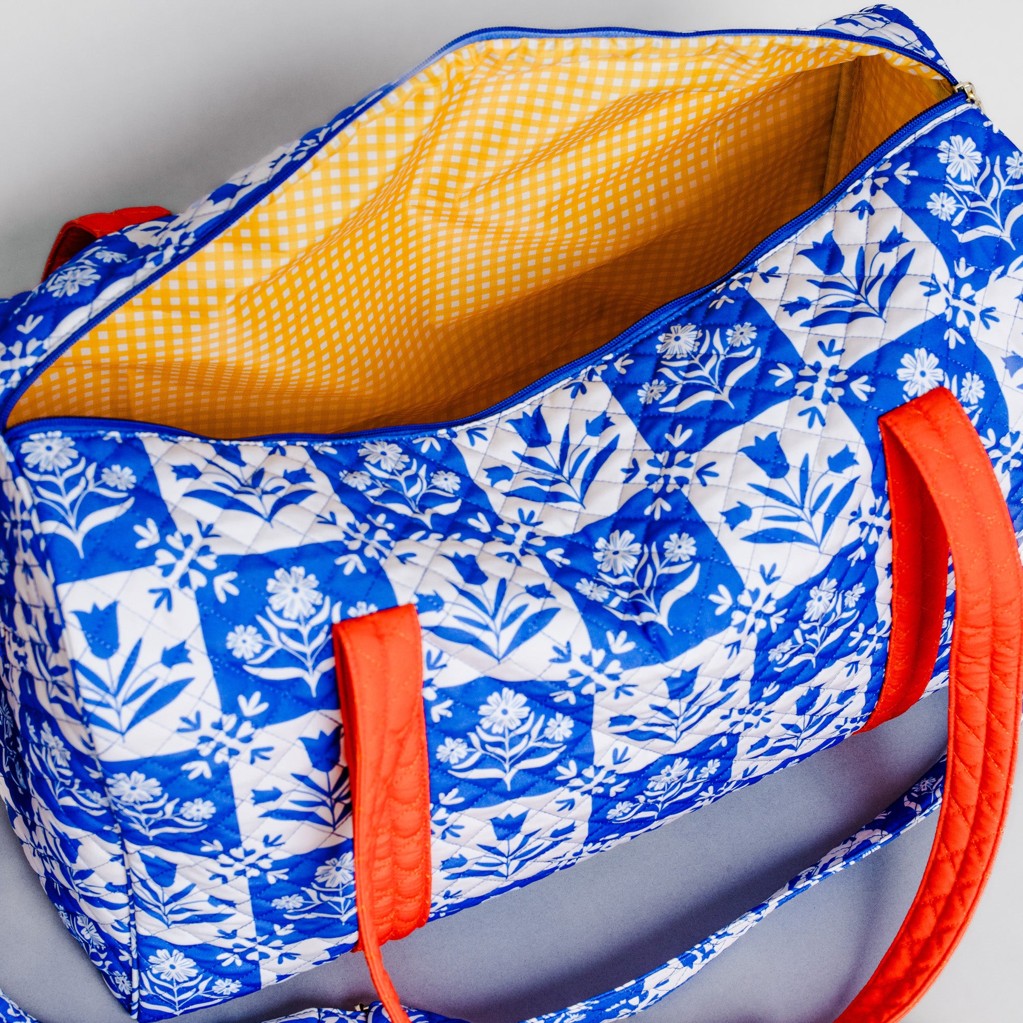 A quilted tote bag with a blue and white floral block-print pattern, a yellow gingham lining, and bright orange handles, shown on a neutral gray background.
