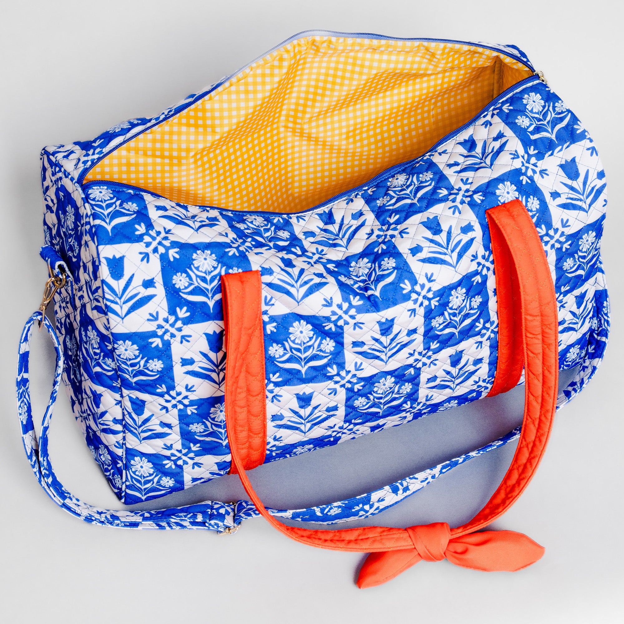 A blue and white patterned duffel bag with a bright yellow gingham interior and orange handles, shown on a light gray background.