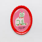 An oval enamel pin featuring a white cat with green details on a pink background, surrounded by a bright red scalloped border with gold trim.