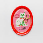 An oval enamel pin holder with a bright red scalloped border, featuring a pink center with a white cat illustration and gold-tone jewelry displayed inside, set against a clean white background.