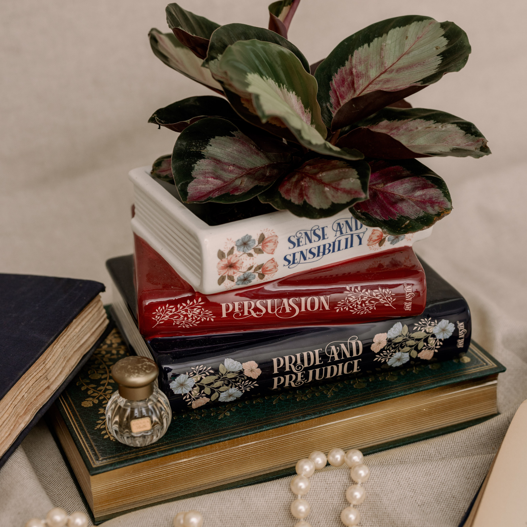 Stack of books with decorative covers, a plant, and a small bottle on a textured surface.