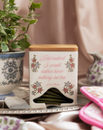 Tea bag container with floral design and text on a table with a plant and pink item in the background